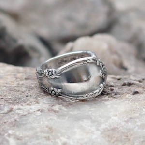 May include: A silver ring made from a spoon, with a floral pattern around the rim. The ring has a broad band and a shiny, reflective surface. The ring is displayed against a neutral, rocky backdrop, emphasising its unique, handmade quality.