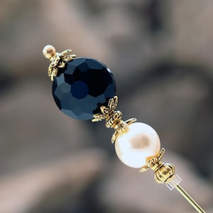 A classically styled Ladies hat pin featuring a  jet black faceted glass coin bead set above a premium faux glass cream pearl.  It is accented with rich gold plated bead caps and spacer beads.