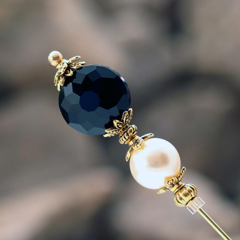 A classically styled Ladies hat pin featuring a  jet black faceted glass coin bead set above a premium faux glass cream pearl.  It is accented with rich gold plated bead caps and spacer beads.