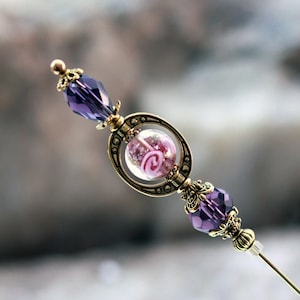 A hat pin with a violet lamp-work style glass bead set inside a gold plate bead cage.  The core of this bead is violet purple with a pink stylized flower and set between a teardrop and a rondelle purple fine cut crystals and accented with gold plate
