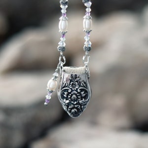 May include: A silver-toned metal pendant featuring an embossed floral pattern, suspended from a chain adorned with white oval beads, small purple accents, and clear faceted crystals. The pendant has an open top with a visible cream-coloured fabric lining.