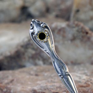 May include: A silver spoon with an ornate handle. The end of the handle has a round hole with a black centre and a ring of clear rhinestones.