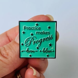May include: A teal blue enamel pin with a black border and black text that reads "Practice makes Progress - dream - believe" with stars around the text.