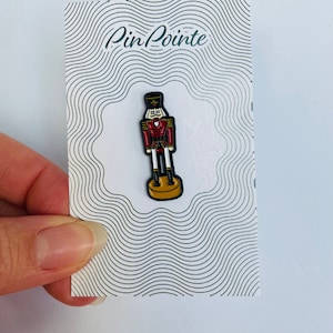 May include: A red and white enamel pin of a nutcracker soldier with a black hat and gold trim. The pin is on a white card with the text "Pin Pointe" in green.