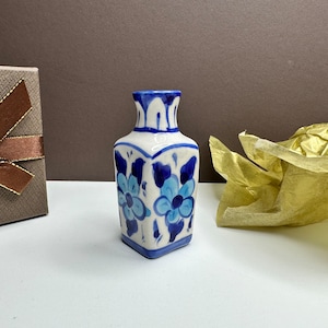May include: A small, white ceramic vase with a square base and a narrow neck. The vase is decorated with blue floral patterns and accents. A brown gift box with a ribbon is to the left, and gold tissue paper is to the right.