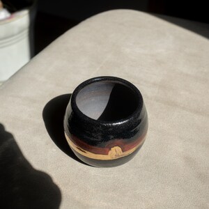 May include: A handcrafted ceramic bowl. The exterior is black, with a band of red and gold near the base. The interior is a lighter brown. The bowl is round and has a smooth, glossy finish.