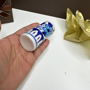 May include: A cylindrical ceramic object featuring a hand-painted blue and white floral pattern. The design includes stylized blue flowers and leaf motifs against a white background, with a solid white base and a smooth, glossy finish throughout.
