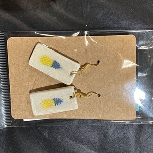 May include: A pair of rectangular earrings featuring a white speckled base with a yellow and blue pineapple graphic. The earrings are attached to gold-toned metal hooks and are presented on a brown cardboard display card inside a clear plastic bag.