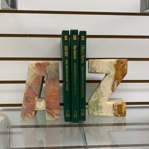 May include: Stone bookends in the shape of the letters "A" and "Z", with a marbled effect in beige, brown, and pink. Three green books are positioned between the bookends. The bookends are a decorative item.