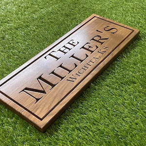 May include: A rectangular brown wooden sign with a smooth finish and a carved border. The text "THE MILLER'S" is engraved in large capital letters, with "WICHITA, KS" engraved in smaller capital letters below it on the right side.