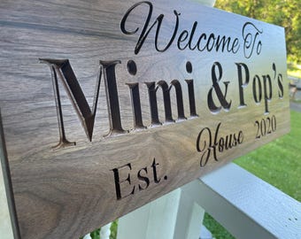 May include: A rectangular dark brown wooden sign with a visible grain pattern. The sign features deeply carved black lettering that reads Welcome To Mimi & Pop's House Est. 2020, with the text arranged in a mix of script and serif fonts.