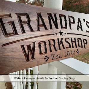 May include: A rectangular wooden sign with the words "GRANDPA'S WORKSHOP" carved into it. The sign includes the text "Est. 2020" and two stars. The sign is made of walnut and is designed for indoor display.