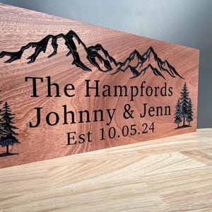 May include: A rectangular wooden sign with a natural reddish-brown grain featuring a black engraved mountain range at the top and pine trees on the sides. The text reads: The Hampfords, Johnny & Jenn, Est 10.05.24, all in black lettering.