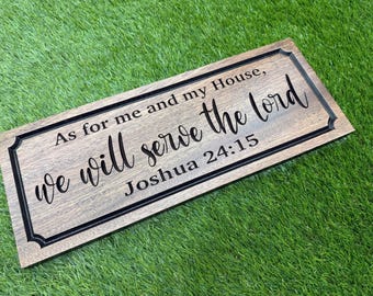 May include: A rectangular wooden sign with a dark brown finish and a beveled edge. The text "As for me and my House, we will serve the lord Joshua 24:15" is engraved in black, featuring a mix of serif and cursive script lettering.