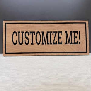 May include: A rectangular wooden sign with a light brown wood grain finish. The sign features a black border and the text "CUSTOMIZE ME!" in bold, black, sans-serif capital letters centered on the front surface of the wooden board.