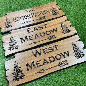 May include: Three wooden signs with black text and tree illustrations. The signs read "The Bottom Pasture", "East Meadow", and "West Meadow", each with an arrow. The signs have a rustic, natural wood grain appearance.