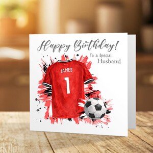 K&ouml;nnte beinhalten: Geburtstagskarte mit einem roten Fu&szlig;balltrikot mit dem Namen "JAMES" und der Nummer "1". Ein Fu&szlig;ball liegt neben dem Trikot. Der Text der Karte lautet "Happy Birthday! To a Special Husband".