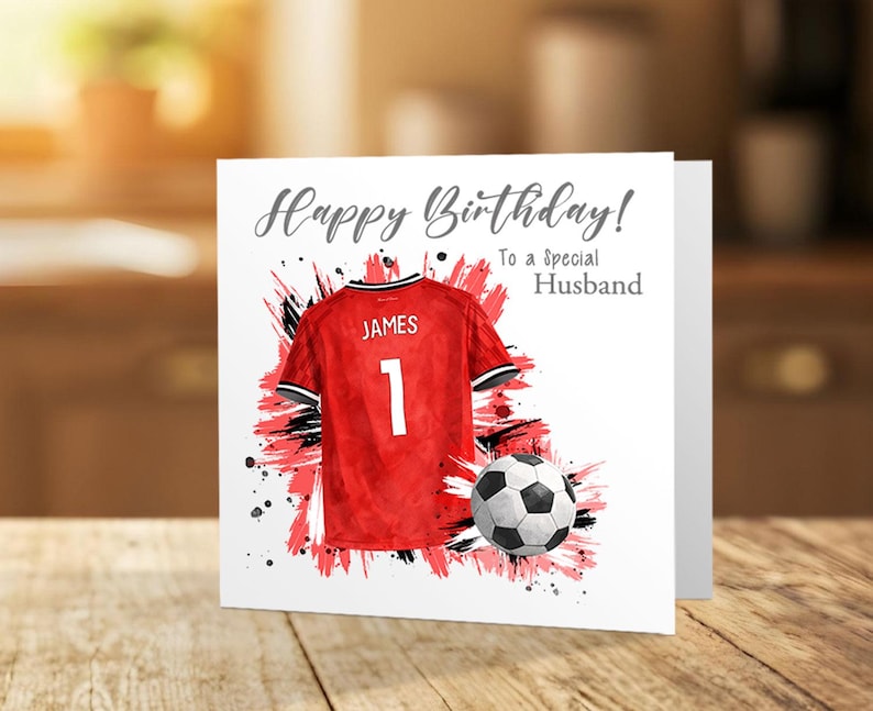 K&ouml;nnte beinhalten: Geburtstagskarte mit einem roten Fu&szlig;balltrikot mit dem Namen "JAMES" und der Nummer "1". Ein Fu&szlig;ball liegt neben dem Trikot. Der Text der Karte lautet "Happy Birthday! To a Special Husband".