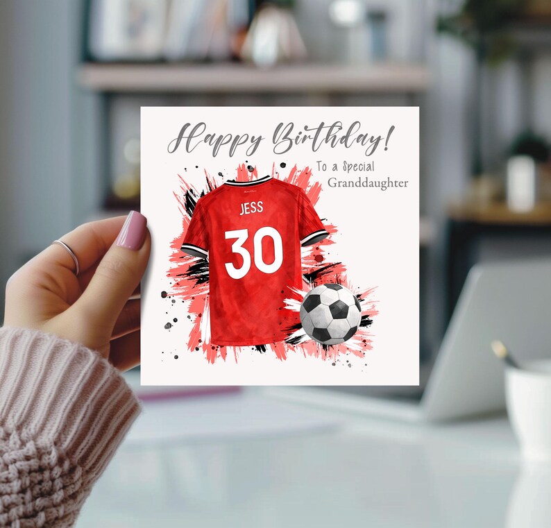 K&ouml;nnte beinhalten: Geburtstagskarte mit dem Text "Happy Birthday! To a Special Granddaughter." Die Karte zeigt ein rotes Fu&szlig;balltrikot mit dem Namen "JESS" und der Nummer "30" sowie einen Fu&szlig;ball. Der Hintergrund hat rote und schwarze Farbspritzer.