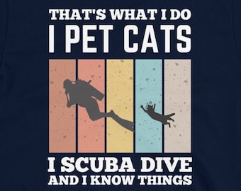 Op de afbeelding: Donkerblauw T-shirt met een retro-stijl grafische afbeelding. De afbeelding toont een duiker met een kat, en de tekst "That's what I do I pet cats I scuba dive and I know things".