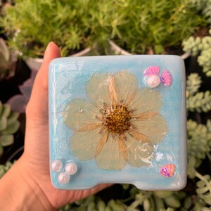 Puede incluir: Una bandeja cuadrada de resina azul claro con una flor prensada y detalles de perlas. La flor es blanca con un centro amarillo. La bandeja tiene un acabado brillante.