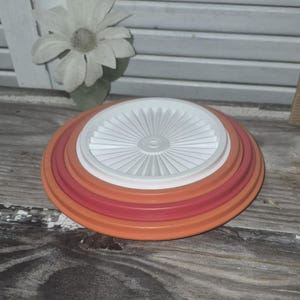 May include: A stack of three circular plastic lids in descending sizes. The bottom lid is orange, the middle lid is a deep reddish-pink, and the top lid is white with a sunburst pattern of radiating ridges extending from the center to the edge.