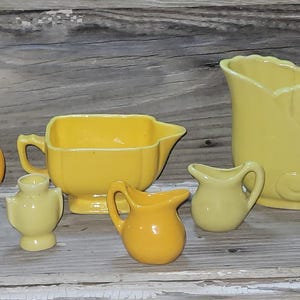 May include: A collection of vintage yellow ceramic serving pieces, including a pitcher, creamer, vase, and small decorative items. The pieces are arranged on a weathered wooden surface, with yellow flowers in the background.