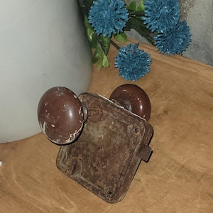 May include: An antique door lock set with two brown, round knobs and a rectangular, rusty metal plate. The lock is set on a wooden surface, with blue artificial flowers and a white vase in the background.