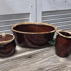 May include: A brown ceramic kitchen set featuring a large bowl, a small lidded jar, and a pitcher. The set has a glossy finish with a lighter brown rim and a rustic, vintage aesthetic. The pieces are arranged on a weathered wooden surface.