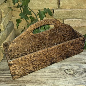 May include: A rectangular wooden caddy with a dark brown stained finish and a prominent wood grain pattern. The caddy features a high central divider that includes an integrated handle cutout for carrying, providing two open storage compartments.
