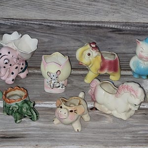 May include: A collection of vintage ceramic animal planters in various colors. The planters include elephants, pigs, a horse, and a rabbit. The planters are arranged on a weathered wooden surface.
