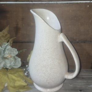 May include: A cream-colored ceramic pitcher with a speckled brown pattern throughout its surface. The pitcher features a curved handle on the right side, a wide base, and a tall, elongated spout designed for pouring liquids with ease.