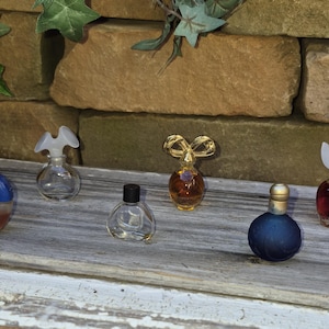 May include: Six miniature perfume bottles of varying shapes and colors. One has a blue cap with the word Navy, one is clear with a white flower cap, one is clear with a black cap, one is gold with a bow, one is dark blue, and one is red with a white flower cap.