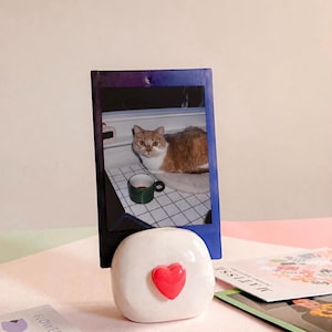 May include: A white ceramic photo holder with a glossy finish and a small, raised red heart on the front. The holder supports a vertical instant photograph featuring an orange and white cat sitting next to a green mug on a white grid surface.