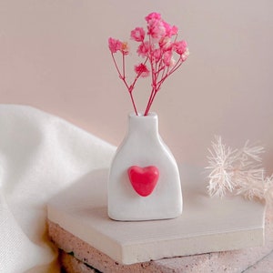 May include: A white ceramic vase with a heart-shaped design on the front. The vase is filled with pink dried flowers and is sitting on a stack of beige tiles.