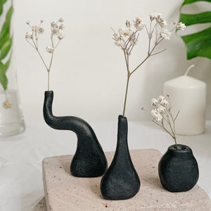 May include: Three black ceramic vases of different shapes and sizes, each holding a sprig of white baby's breath flowers.