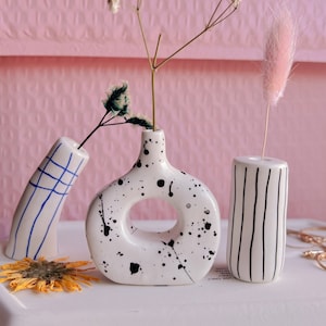 May include: Three white ceramic vases with different designs. The vase on the left has blue and white stripes. The vase in the center has black paint splatters and a ring cutout. The vase on the right has black vertical stripes.