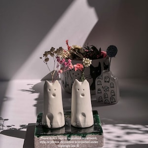 Puede incluir: Dos jarrones de cerámica blanca con forma de gato con flores secas. Los jarrones tienen una superficie texturizada y están sobre una baldosa verde y blanca. El texto "@Clayousomuch" es visible en la imagen.
