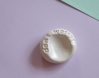 May include: A small, white ceramic dish with the words "don't worry" embossed on the surface. The dish has a smooth, glossy finish and is set against a pastel purple background with a mint green accent.