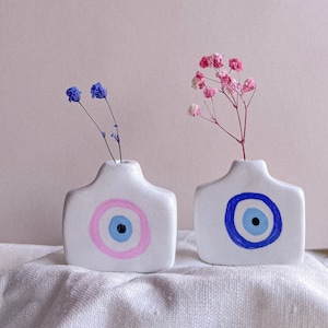 May include: Two white ceramic vases with blue and pink evil eye designs. The vase on the left has a pink eye and the vase on the right has a blue eye. Both vases have dried flowers.