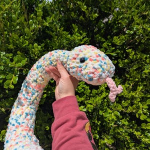 May include: A handmade, plush snake toy in white with colorful speckles of blue, yellow, and pink. The snake has a pink tongue and black eyes. The toy is held up against a backdrop of green foliage.