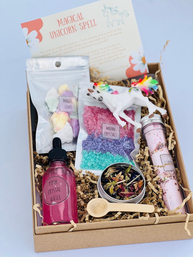 May include: A brown box filled with unicorn-themed bath products. The box contains bath salts, bath bombs, a unicorn figurine, a bottle of pink crystal water, a small tin of dried flowers, and a bath scrub. The box also includes a small wooden spoon and a note that reads "Magical Unicorn Spell".