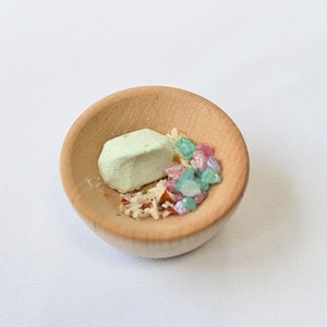 May include: A small wooden bowl filled with green, pink, and white candy. The candy is arranged in a small pile in the center of the bowl.