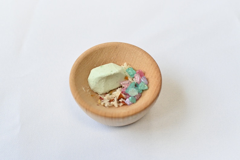 May include: A small wooden bowl filled with green, pink, and white candy. The candy is arranged in a small pile in the center of the bowl.