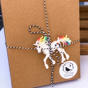 May include: A brown cardboard box wrapped with black and white twine and tied with a rainbow unicorn figurine. The box has a white sticker with a black bird and the text "LITTLE BIRD handcrafted play".
