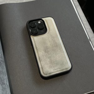 May include: A smartphone case with a black frame and a textured, light beige, stone-like back panel. The case features a triple-lens camera cutout at the top left and the word bluestone printed in small, dark letters near the bottom edge.