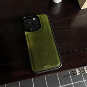 May include: A smartphone case featuring a textured olive green leather back panel with a black frame and camera housing. The word bluestone is printed in small gold letters at the bottom of the green panel. The case has a rectangular shape.