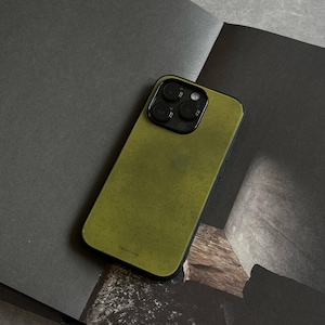 May include: Olive green phone case with a black border. The case has a textured appearance and a camera module with three lenses. The phone case is resting on a dark surface.