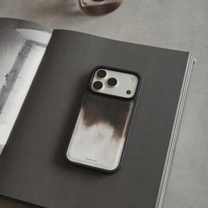 May include: A black and white iPhone case with a unique design, featuring a black border and a white center with a brown, cloud-like pattern. The case is resting on a dark gray book, with a glass of liquid in the background.