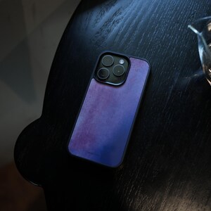 May include: A smartphone case with a purple and blue watercolor design. The case has a black border and a camera cutout. The phone case is resting on a dark wooden surface.